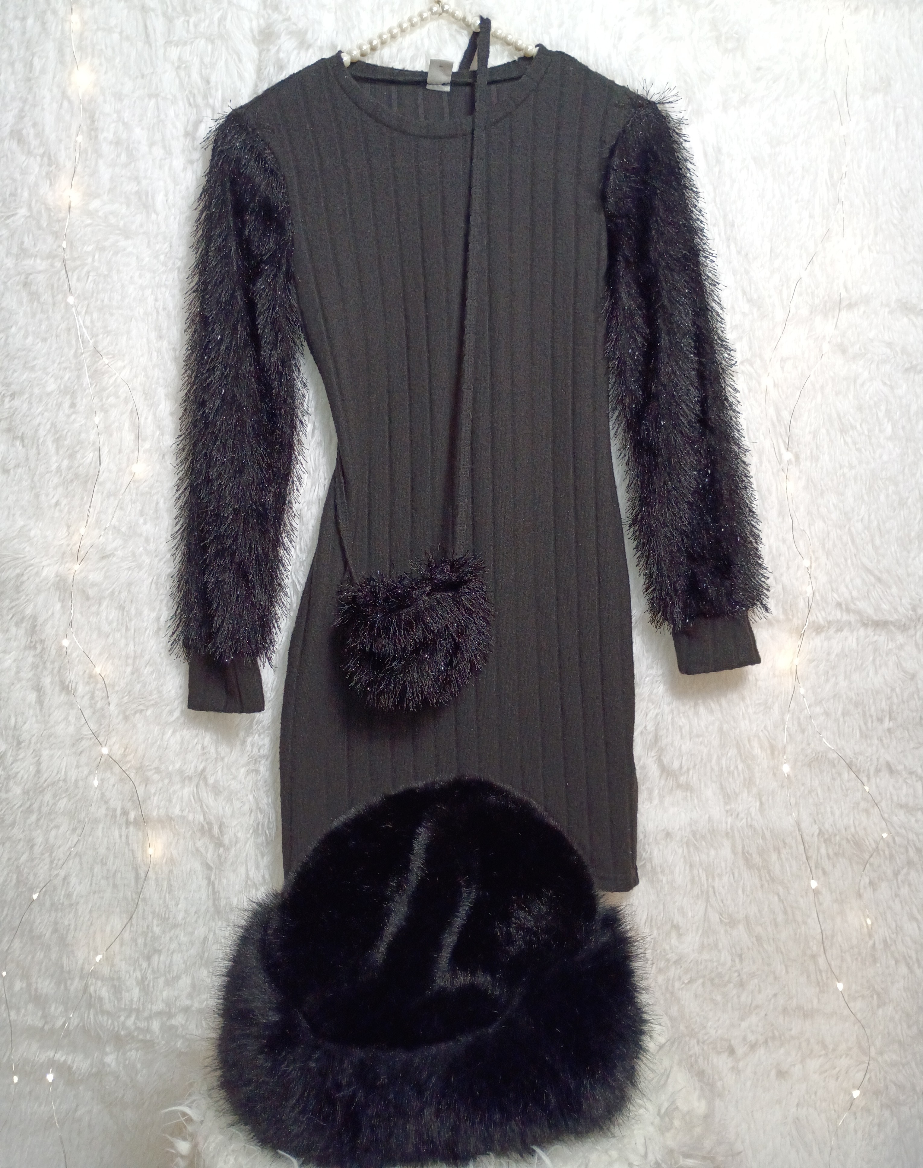 This image features a black long‑sleeved ribbed dress displayed on a soft, light‑colored textured surface. The sleeves and waist include faux‑fur accents, adding warmth and sophistication. A small fuz